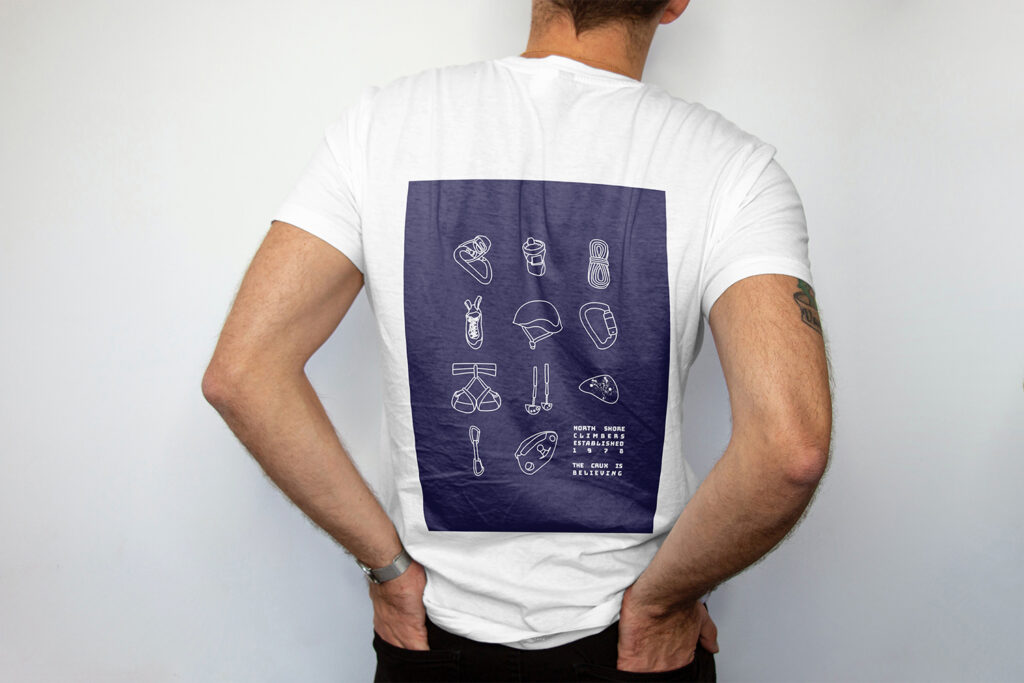 On a t-shirt is a design in a grid, are 11 different climbing equipment pieces (shoes, chalk back, rope, belay device, harness, cams, grigri, etc.) and the words "North Shore Climbers established 1978, the Crux is believing"